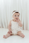 Baby sitting on a white floor wearing a white romper with floral patterns and a matching headband.