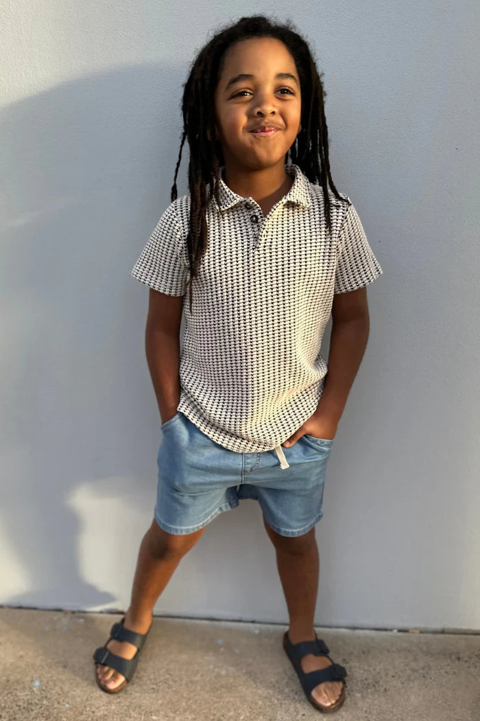 Child wearing a checkered shirt and denim shorts standing against a white wall.