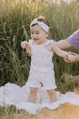 Baby in a white outfit standing in a grassy field with a blurred background