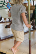 Child in a checkered shirt and beige shorts standing on a wooden floor.