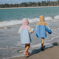 Two people in colorful hooded cloaks walking along a beach with waves and palm trees in the background.