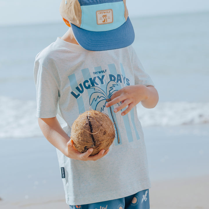 Child wearing a 'Lucky Days' crywolf t-shirt and blue cap holding a coconut on a beach.