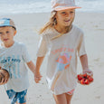 Two children on a beach, one holding a coconut and the other a plate of strawberries, wearing CRYWOLF 'Lucky Days' t-shirts.
