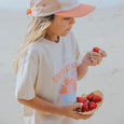 Child wearing a white CRYWOLF t-shirt with colorful graphics and an orange cap, holding strawberries.