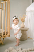 Baby in a white outfit standing next to a crib in a neutral-colored room.