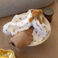 Brown teddy bear wrapped in a white blanket with brown bear patterns on a wooden surface.