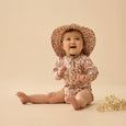 Baby wearing a floral Wilson & Frenchy sunsuit and hat on a beige background