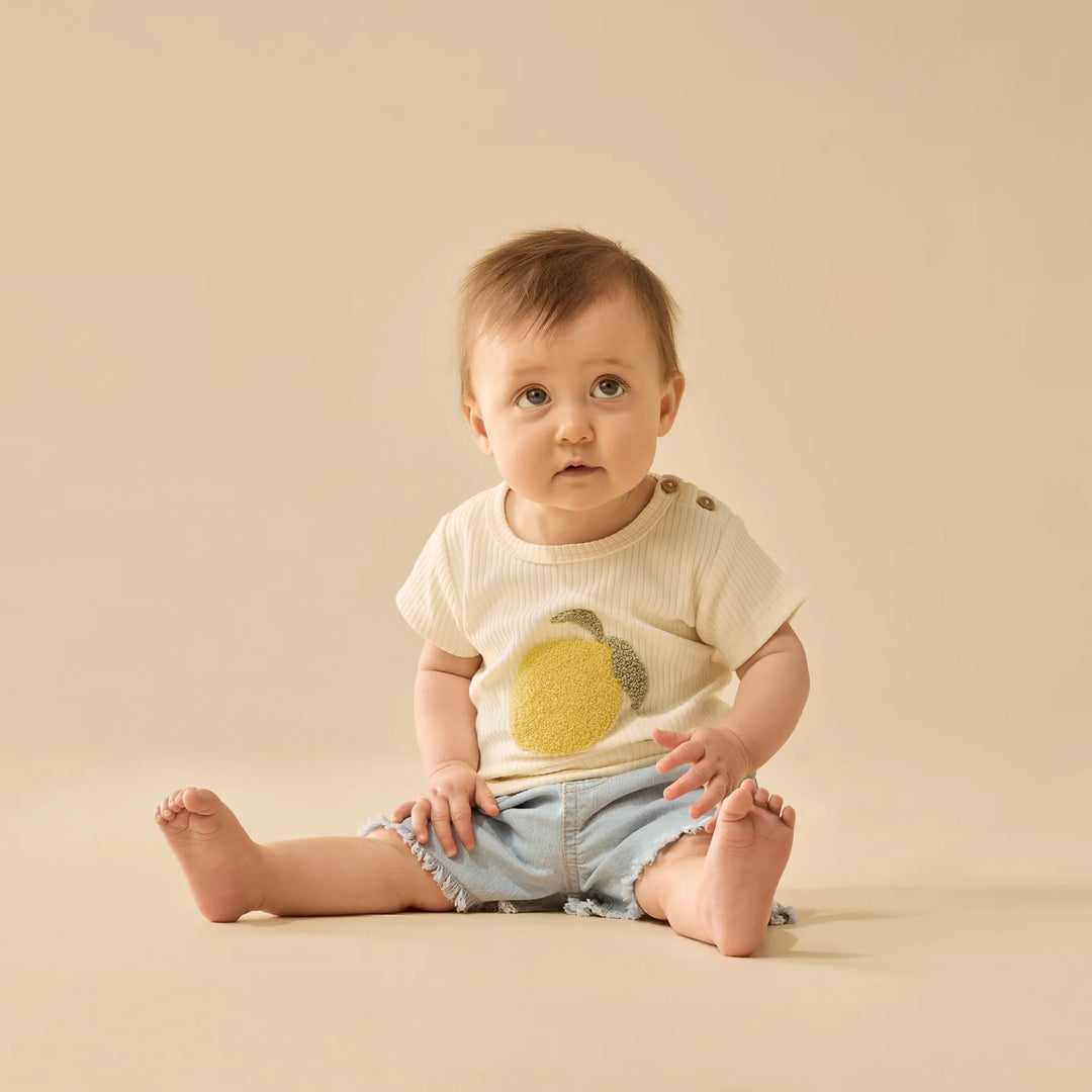Baby sitting on a beige surface wearing a light-colored wilson & frenchy tshirt with a lemon design and denim shorts.
