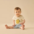 Baby sitting on a beige surface wearing a light-colored wilson & frenchy tshirt with a lemon design and denim shorts.