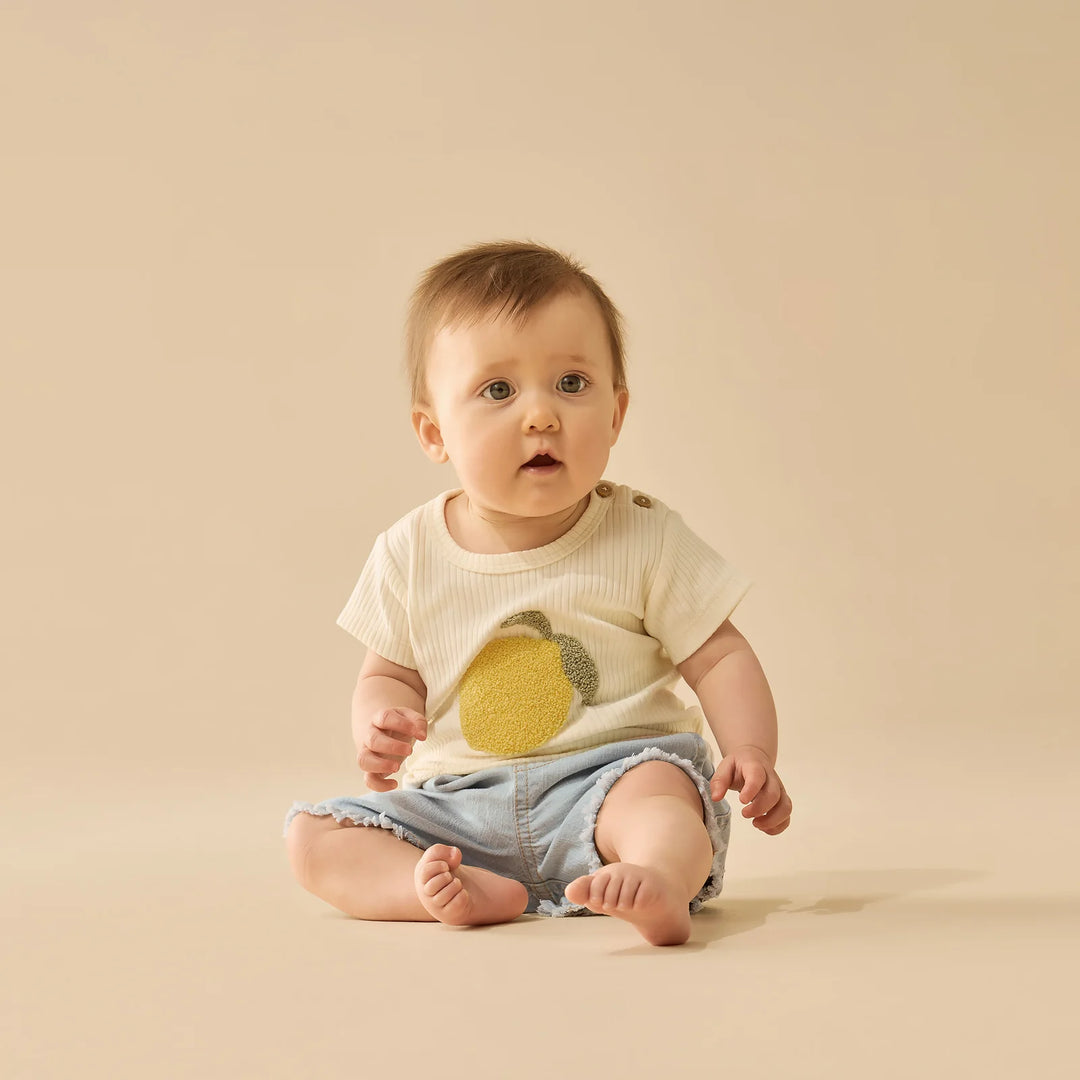 Baby wearing a white Wilson & Frenchy tshirt with a lemon design and denim shorts on a beige background