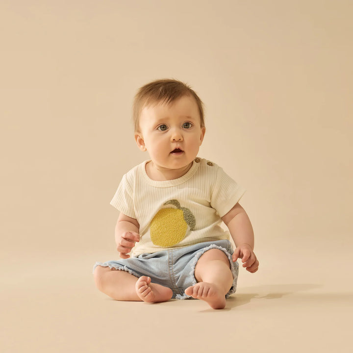 Baby wearing a white Wilson & Frenchy tshirt with a lemon design and denim shorts on a beige background