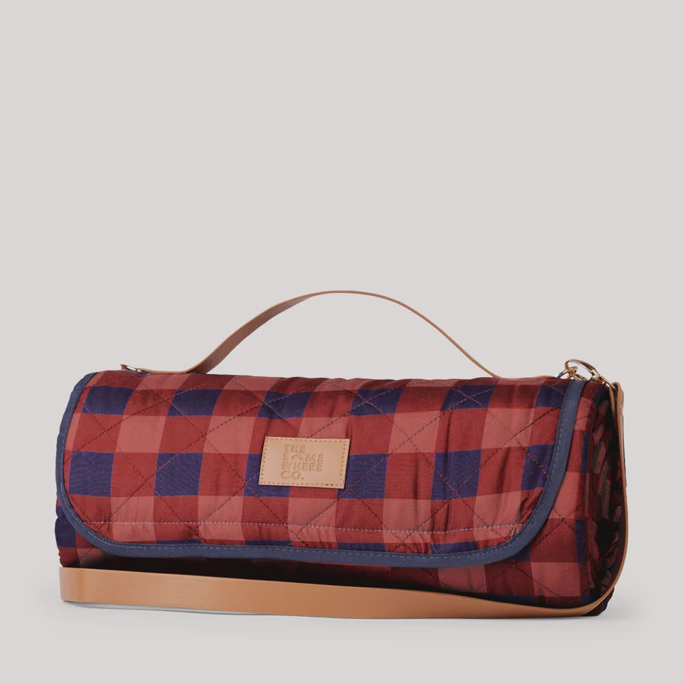 A quilted picnic blanket bag with a red and navy blue buffalo plaid pattern, featuring a tan leather handle and a matching leather brand patch that reads 'Piquadro'.