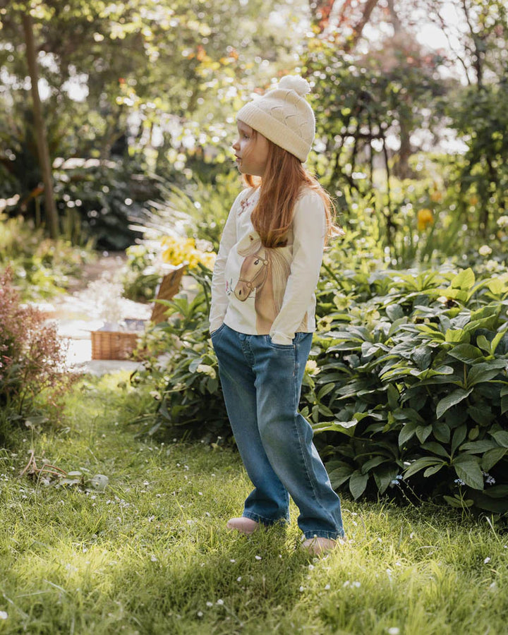 Person standing in a garden wearing a white sweater, blue jeans, and a beige hat.