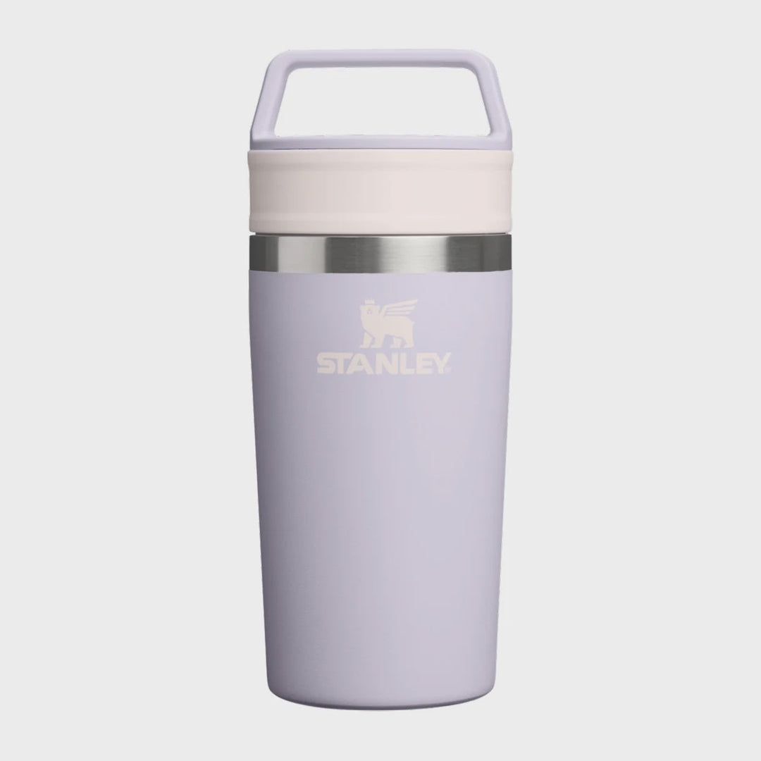 A Stanley travel tumbler in a light lavender purple color, featuring a cream-colored lid with a carrying handle, a silver metallic band, and the Stanley logo printed on the front, set against a solid light gray background.