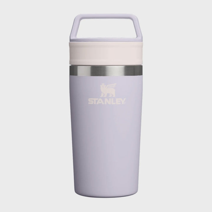 A Stanley travel tumbler in a light lavender purple color, featuring a cream-colored lid with a carrying handle, a silver metallic band, and the Stanley logo printed on the front, set against a solid light gray background.