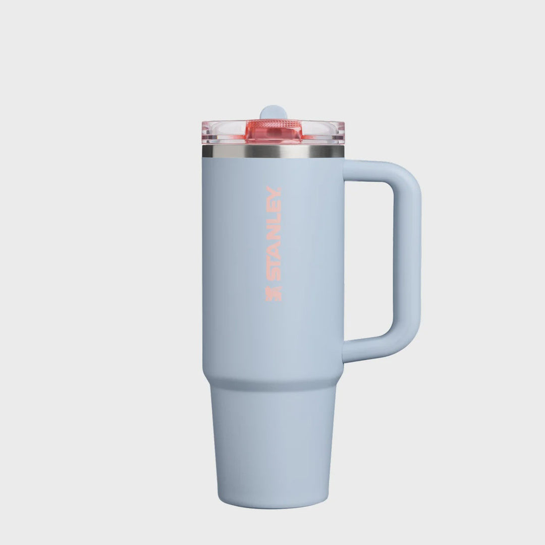 A light blue Stanley travel mug with a handle, a clear lid with a red seal, and the Stanley logo printed vertically in pink on the front, set against a plain white background.