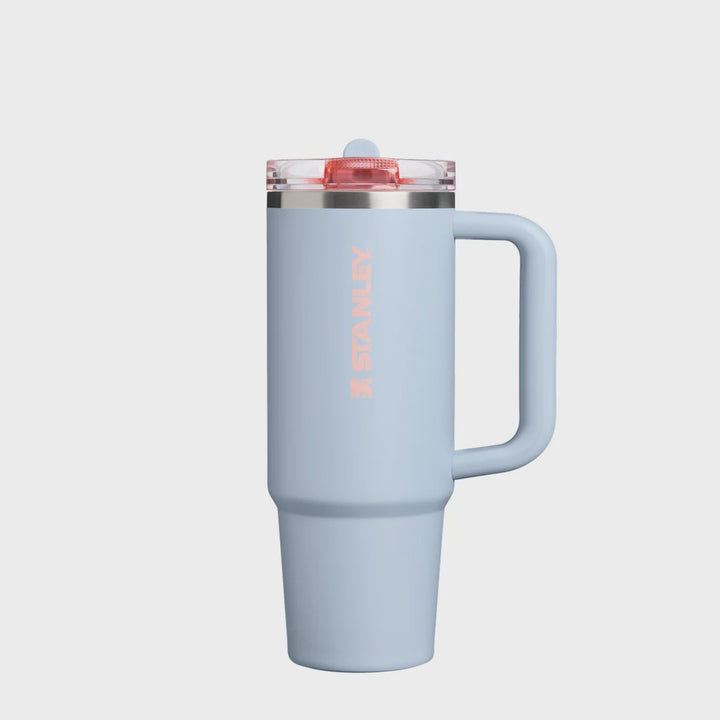A light blue Stanley travel mug with a handle, a clear lid with a red seal, and the Stanley logo printed vertically in pink on the front, set against a plain white background.