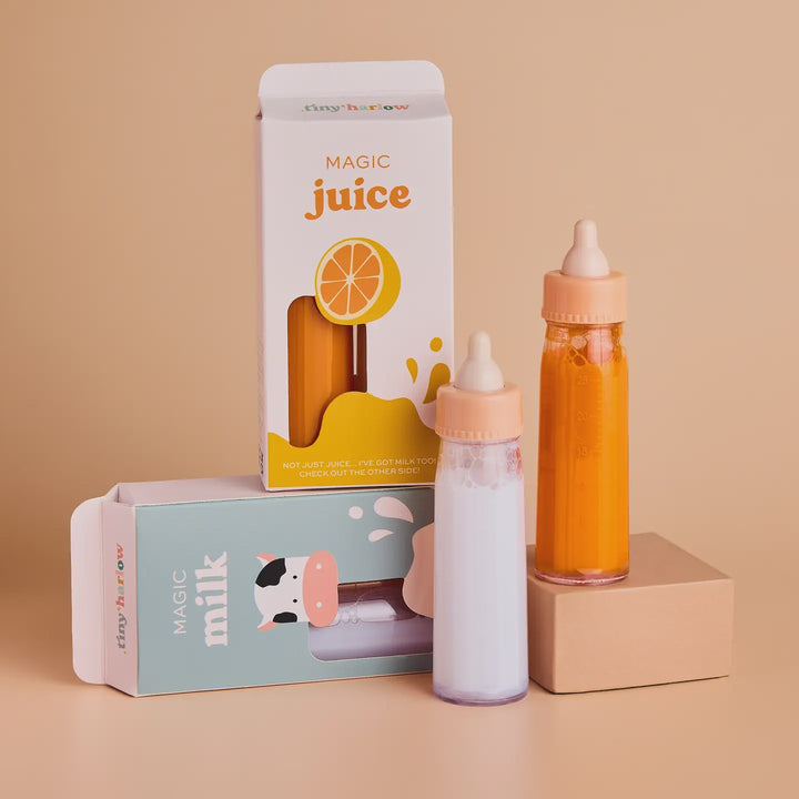 Two baby feeding bottles with peach-colored nipples, one filled with orange juice and the other with white milk, displayed next to their retail packaging boxes on a warm beige background.