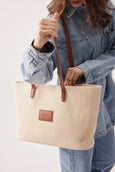 Person holding a beige straw tote bag with brown leather straps against a plain background