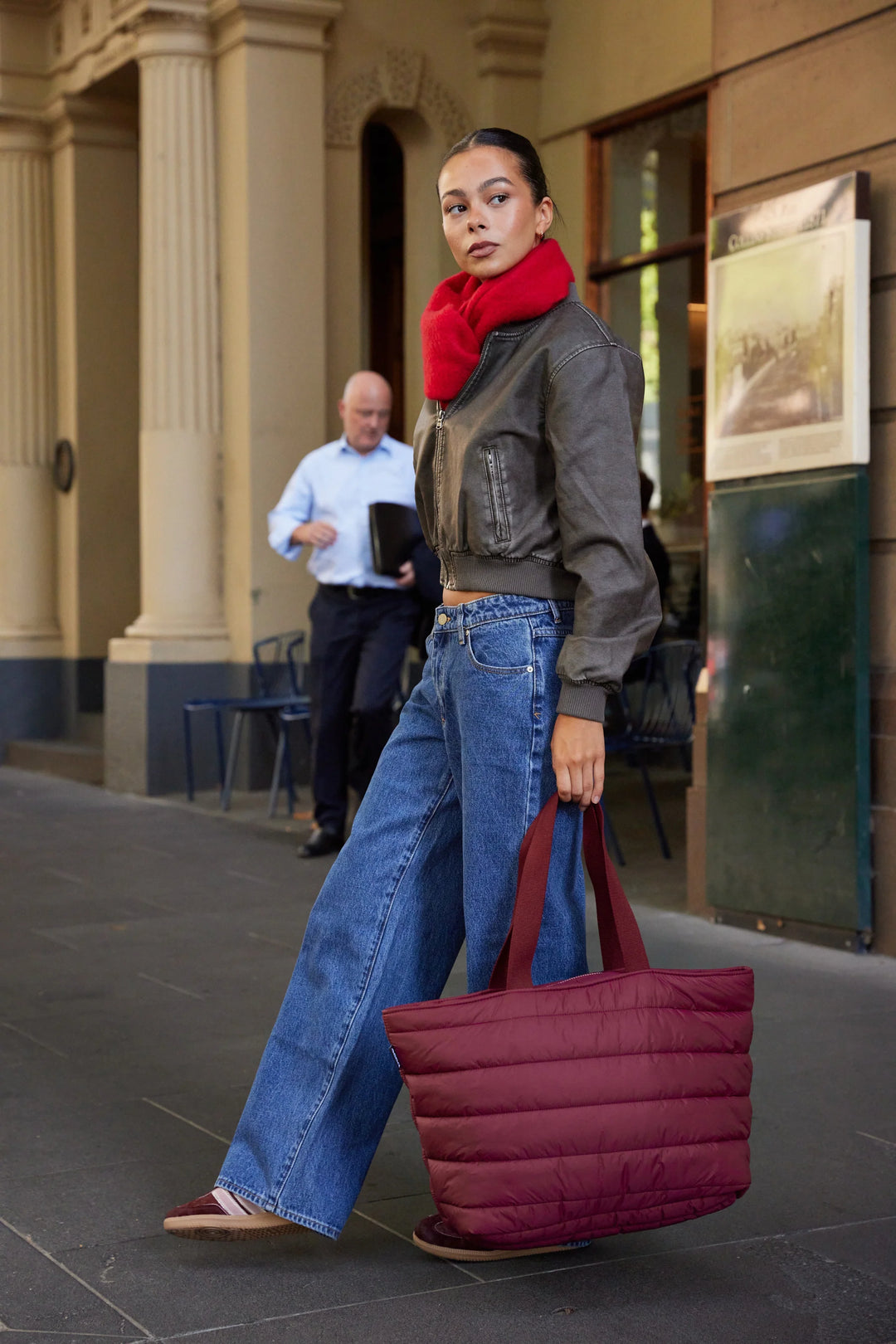 Woman walking outdoors holding a red bag, wearing a red scarf and blue jeans.