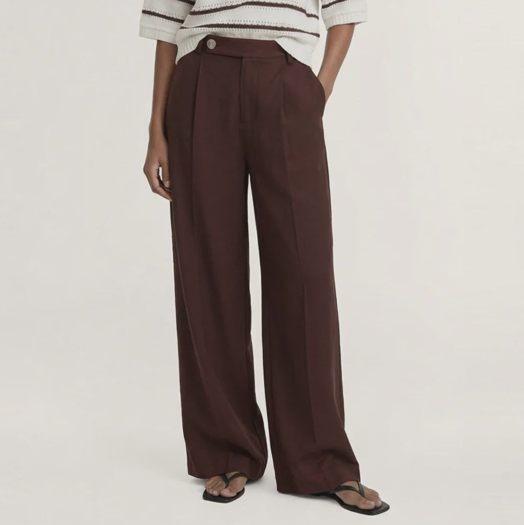 A model stands against a plain light beige studio background wearing a cream knit sweater with dark brown horizontal stripes, paired with dark brown wide-leg trousers and dark sandals.