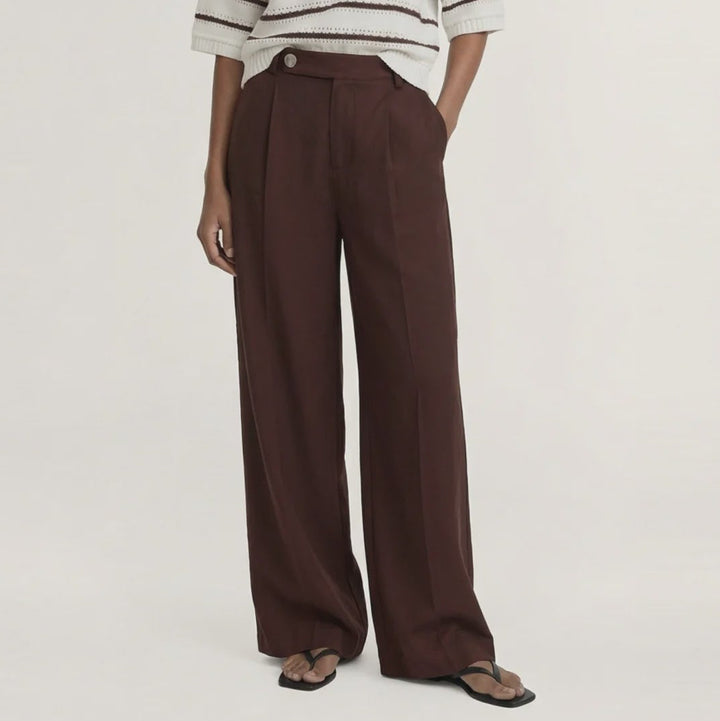 A model stands against a plain light beige studio background wearing a cream knit sweater with dark brown horizontal stripes, paired with dark brown wide-leg trousers and dark sandals.
