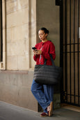 Woman in red jacket and blue jeans standing against a wall, holding a phone and a black bag.