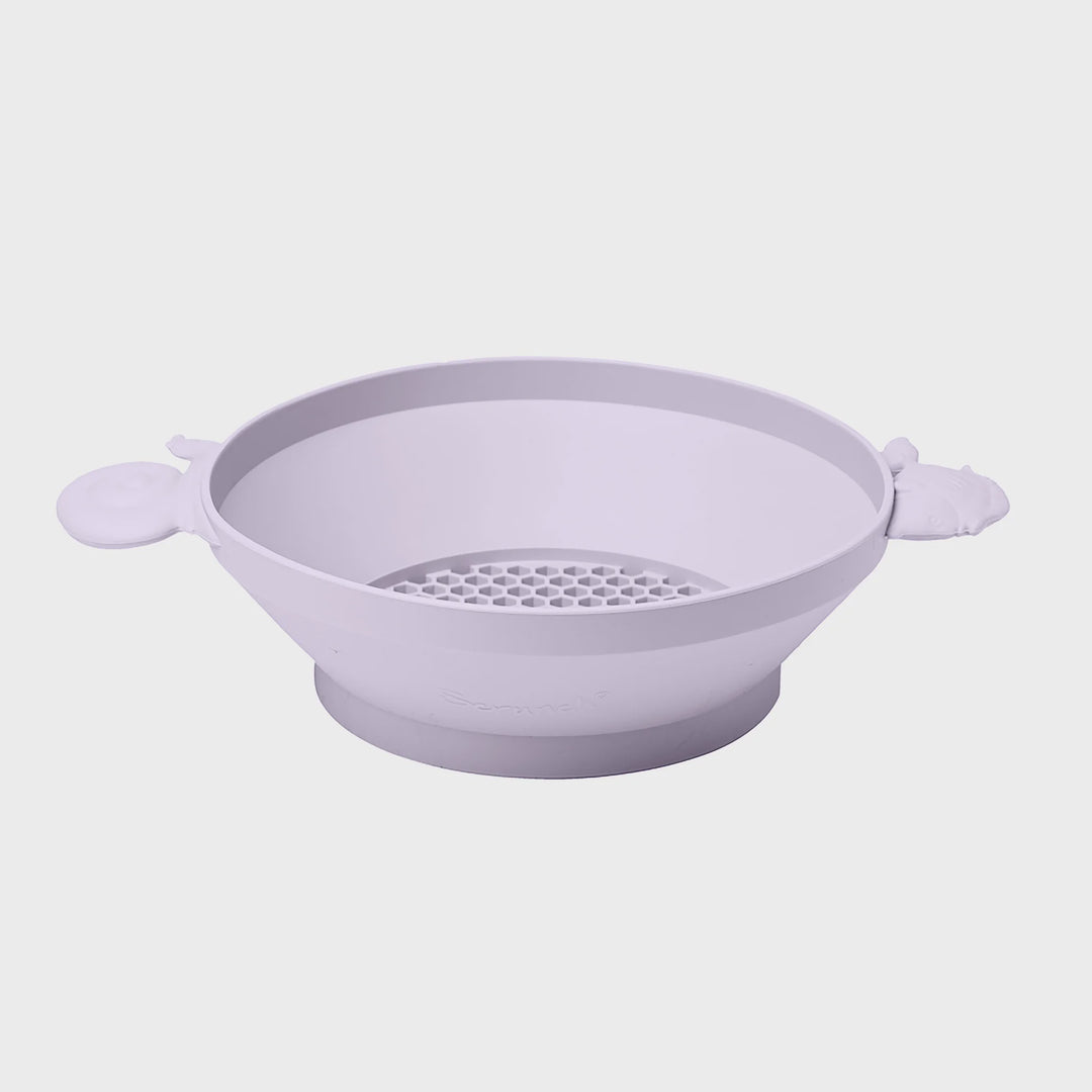 A light lavender silicone baby feeding bowl with integrated animal-shaped side handles and a perforated drain insert, shown on a neutral light gray background.