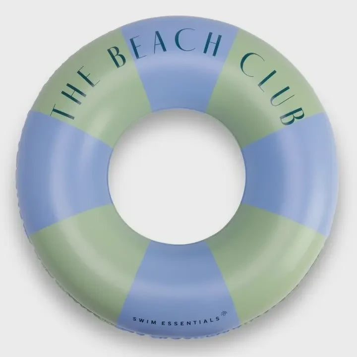 A circular inflatable swim ring with alternating pastel blue and sage green segments, featuring the text 'THE BEACH CLUB' in dark teal lettering and 'SWIM ESSENTIALS' near the bottom, shown on a light gray background.