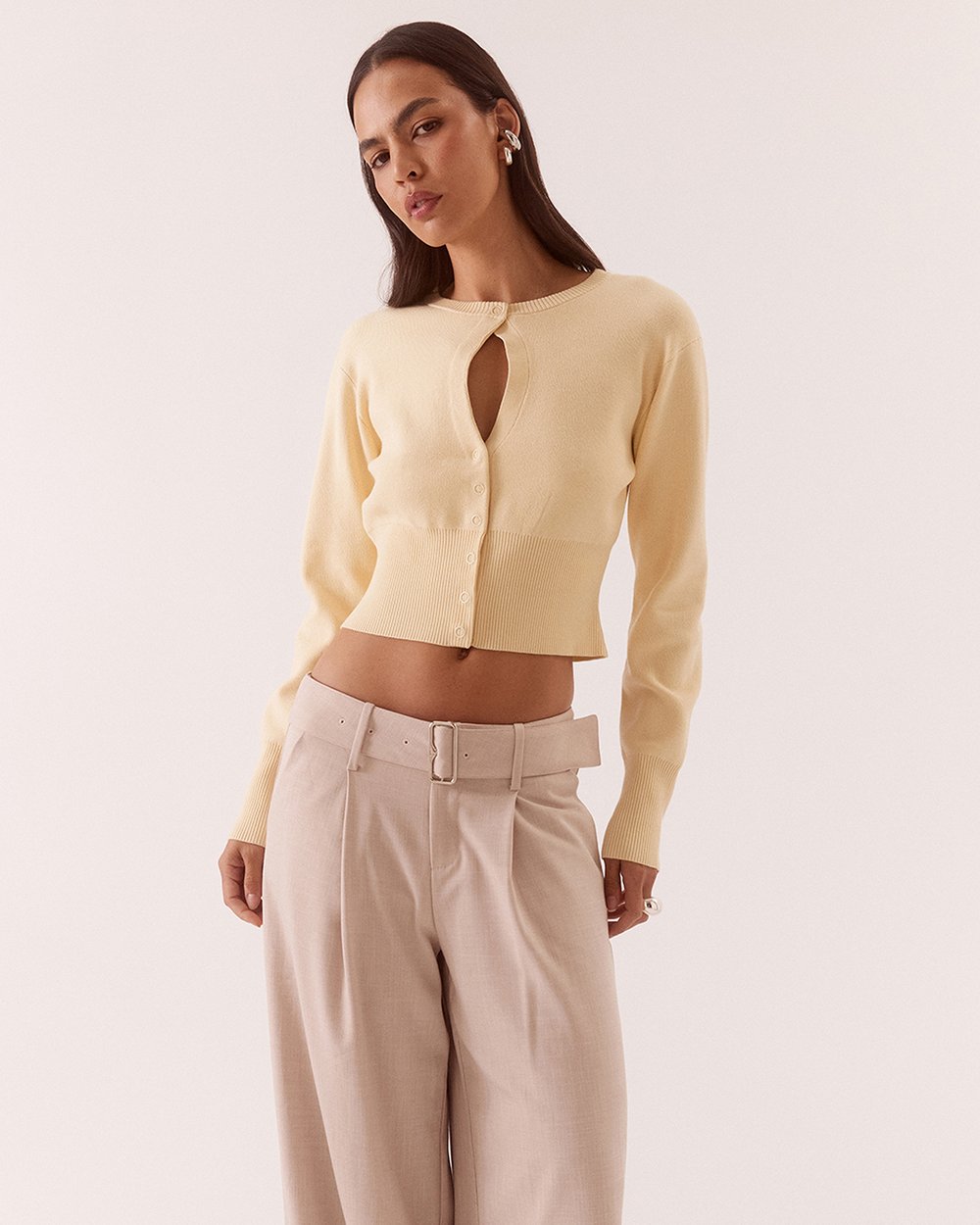 A woman modeling a pale yellow long-sleeve cropped knit top with a keyhole cutout at the neckline, paired with light beige high-waisted trousers, against a solid off-white background.