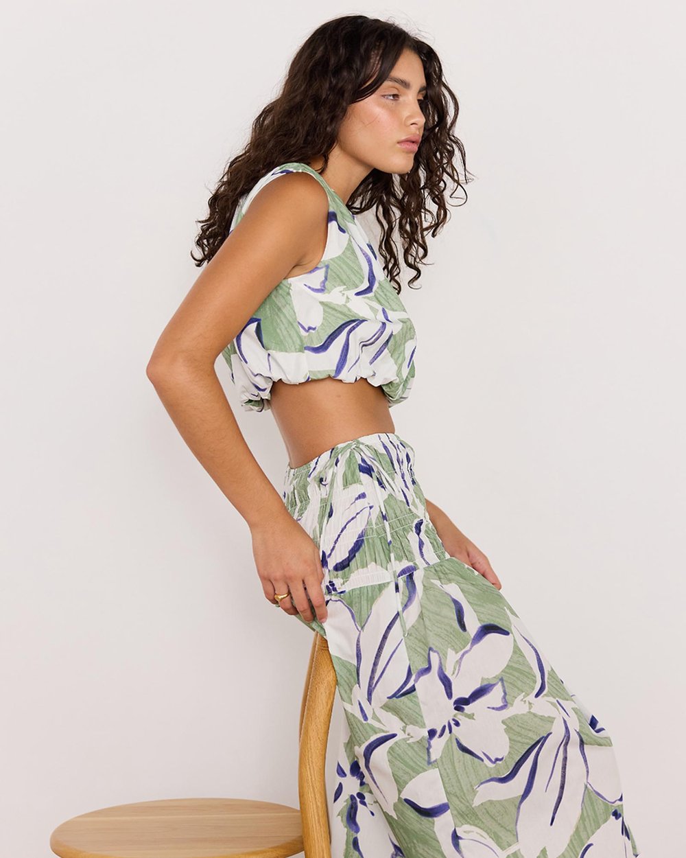 Woman wearing a green and white floral outfit sitting on a wooden stool against a plain background