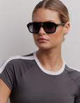 Woman wearing dark sunglasses and a gray athletic shirt with white trim against a plain background