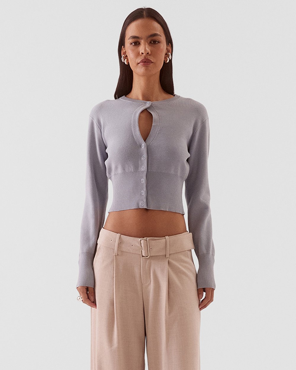 A model wearing a light lavender-gray cropped knit cardigan with a keyhole cutout and beige wide-leg trousers, shot against a solid light gray background.