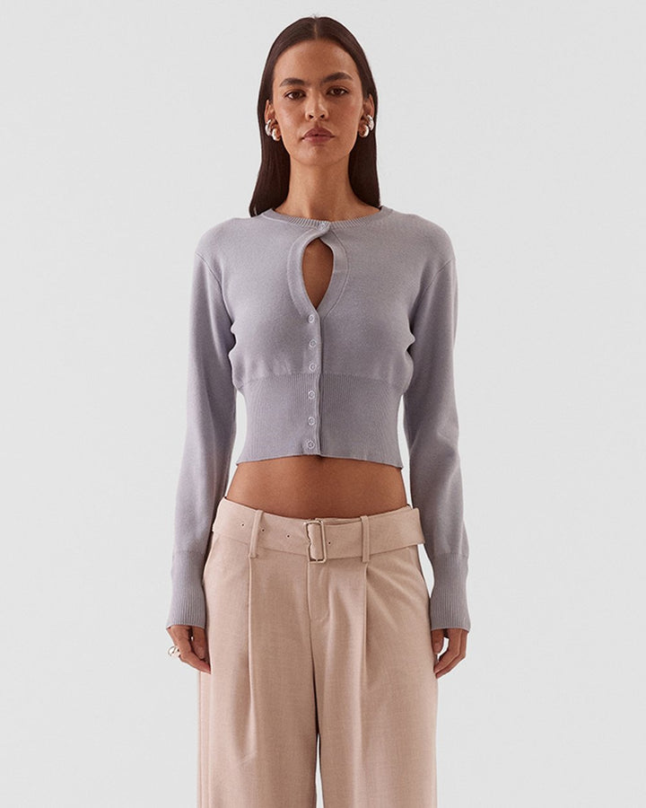 A model wearing a light lavender-gray cropped knit cardigan with a keyhole cutout and beige wide-leg trousers, shot against a solid light gray background.