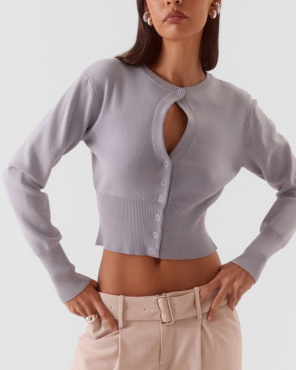 A model wearing a light lavender ribbed knit cardigan with a keyhole cutout and button details, paired with beige trousers, against a solid light gray background.