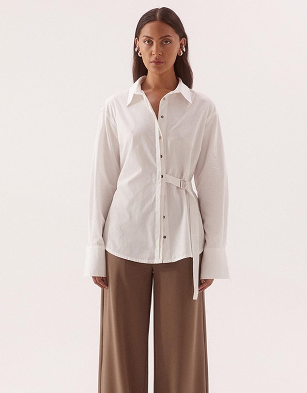 A woman modeling a white long-sleeve button-up shirt with a self-tie belt, paired with brown wide-leg trousers, against a plain light background.