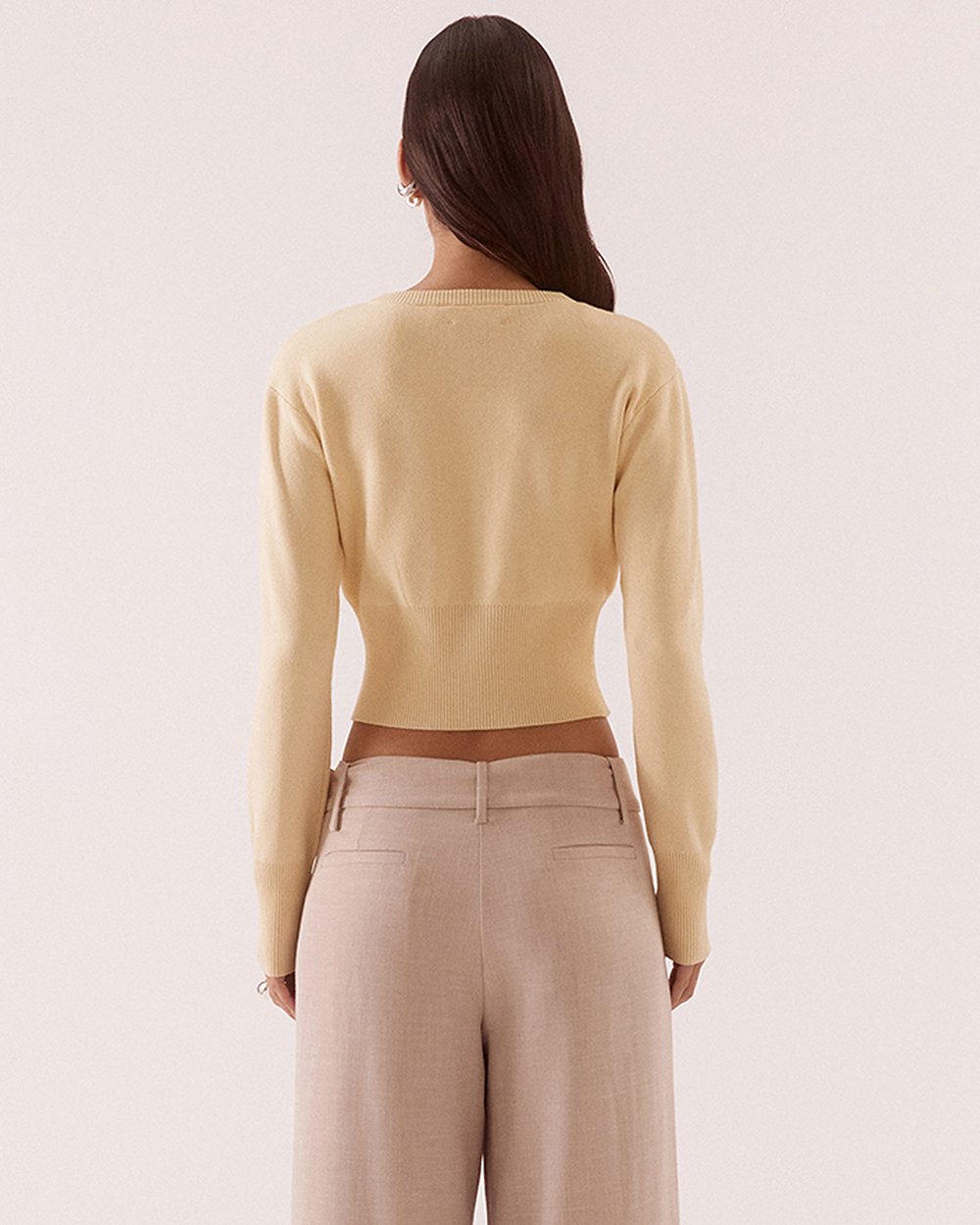 A woman shown from the back wearing a pale yellow long-sleeve cropped knit top and light beige wide-leg trousers against a plain light background.