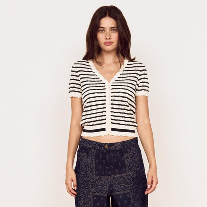 A woman stands against a plain white wall wearing a short-sleeve black and white striped knit cardigan, dark navy wide-leg patterned pants, and brown sandals.