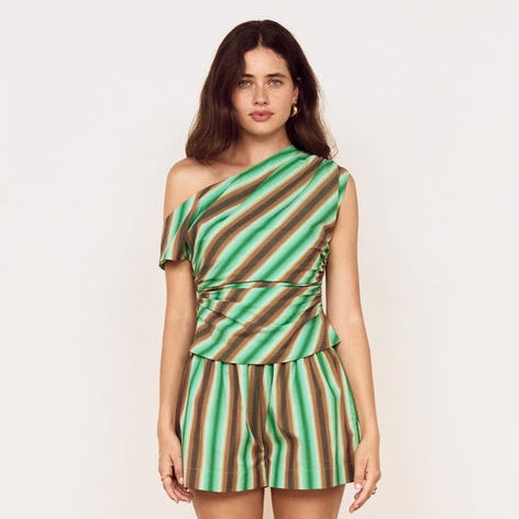A woman modeling a one-shoulder striped romper with green and brown vertical stripes, paired with brown flip-flop sandals, standing against a plain off-white wall on a light wood herringbone floor.