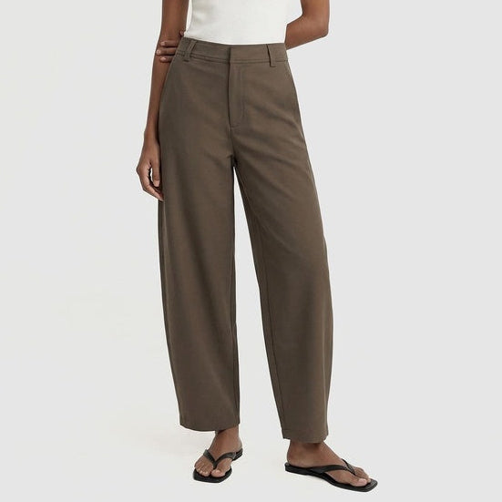 A model wearing high-waisted, wide-leg taupe pants paired with a white tank top and black flip-flops, standing against a light gray studio background.