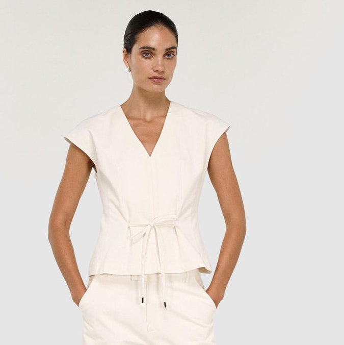 A woman modeling a cream-colored sleeveless wrap-style top with a tie waist and matching wide-leg pants, shown against a plain light gray studio background.