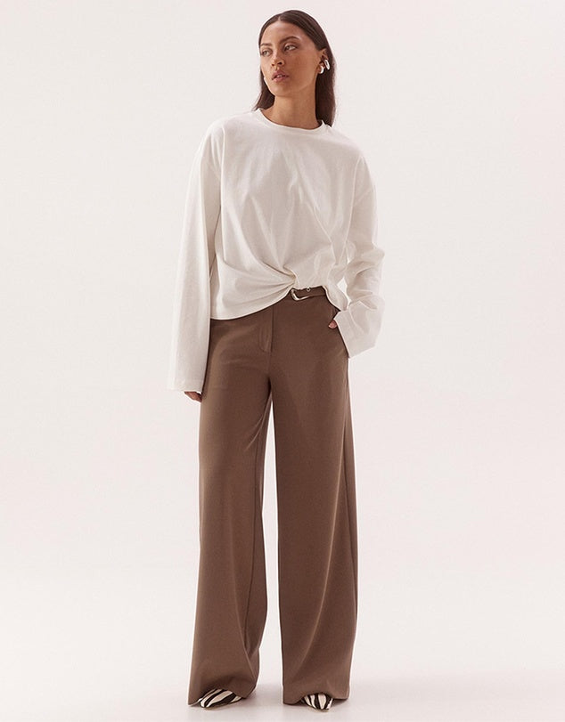 A woman modeling a white long-sleeve top with a twisted hem and brown wide-leg trousers, standing against a plain light background.
