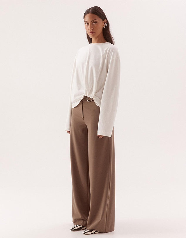 A model is shown from the back wearing a white long-sleeve top with a twisted hem detail and brown wide-leg trousers, set against a plain light background.