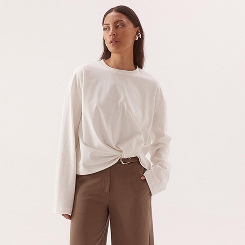 A woman modeling a white long-sleeve top with a twisted hem and brown wide-leg trousers, standing against a plain light background.