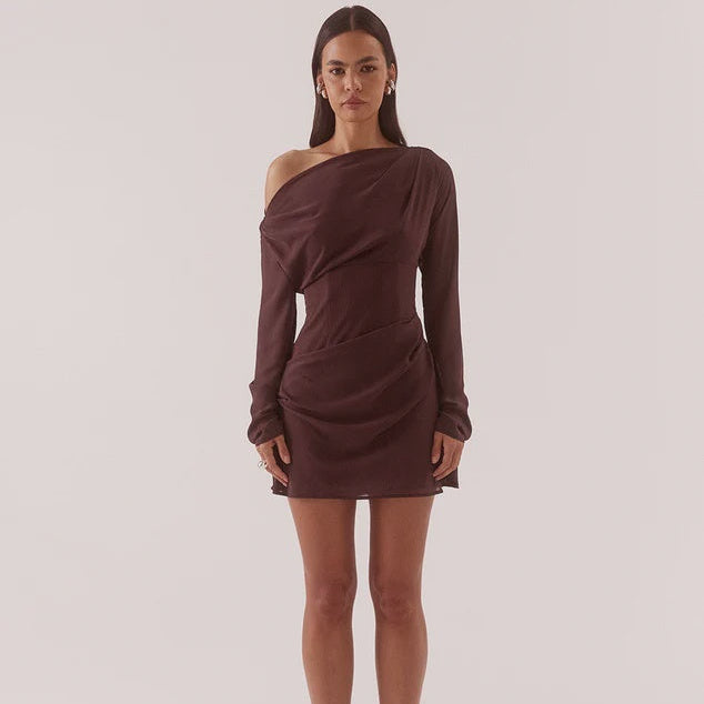 A woman modeling a dark brown, long-sleeved, one-shoulder mini dress with ruched draping details, standing against a solid light beige studio background.