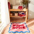 Pink rug with 'Nudie Rudie' text on a wooden floor in a room with a shelf and decorative items.