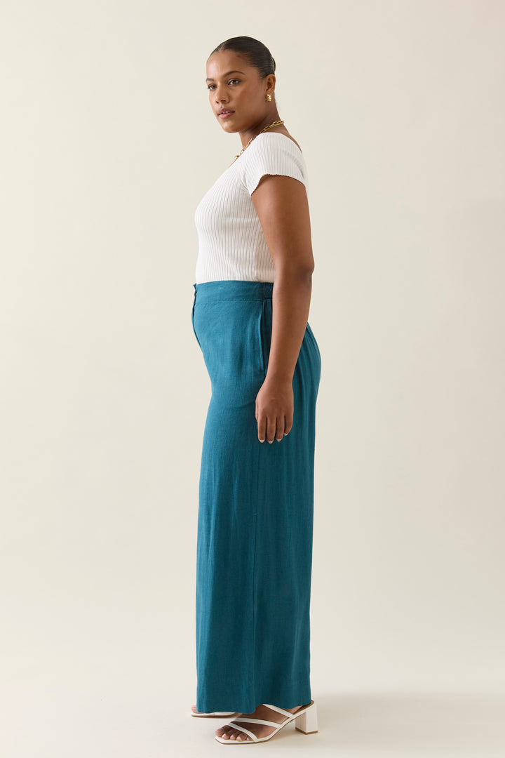 Woman wearing a white top and teal isle of mine pants on a plain background