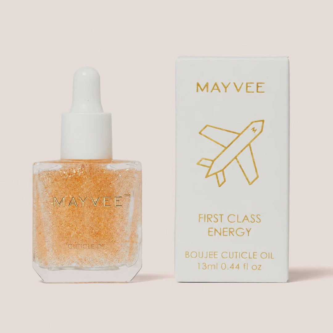 May Vee First Class Energy Bougie Cuticle Oil in a clear square bottle filled with shimmering orange oil, shown next to its white packaging box with gold lettering on a pale pink background.