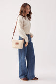 Woman wearing a cream sweater, blue jeans, and brown shoes with a beige handbag on a white background