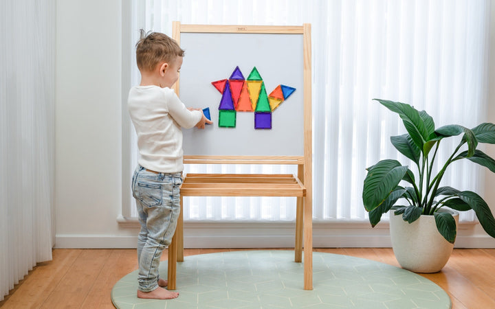 Child playing with connetix colorful magnetic shapes on a wooden easel in a bright room.